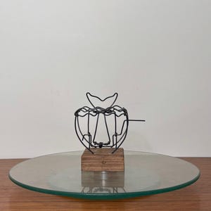 Puede incluir: Una escultura de alambre negro con forma de manzana, montada sobre un pequeño bloque de madera. La escultura se encuentra sobre un plato de cristal redondo y transparente con borde verde, expuesto sobre una superficie de madera.