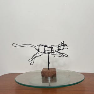 May include: A wire sculpture of a cat in a leaping pose, mounted on a wooden base. The sculpture is made of black wire and is set on a clear glass plate.