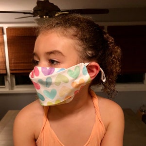 May include: A young person wearing a white face mask with a colorful heart pattern. The mask has elastic ear loops and covers the nose and mouth.