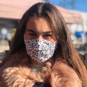 May include: A young person wearing a white face mask with a colourful leopard print pattern. The mask has a light blue stripe across the centre. The person is wearing a brown faux fur jacket.