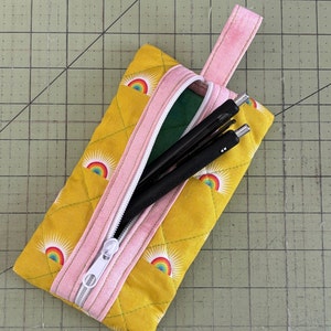 May include: A yellow pencil case with a pink zipper and a pink handle. The case has a repeating pattern of rainbows and suns. The case is open and has black pens inside.