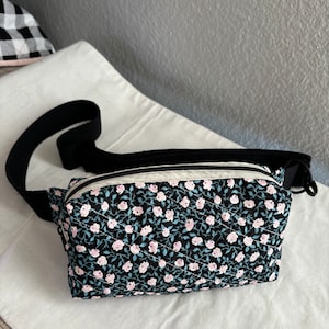 Könnte beinhalten: Eine schwarze Bauchtasche mit einem weißen Reißverschluss und einem Blumendruck mit rosa Rosen auf schwarzem Hintergrund. Die Tasche hat einen verstellbaren schwarzen Gurt.