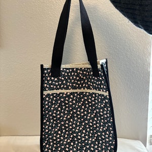 Puede incluir: Una bolsa de mano negra con asas negras y un estampado floral con flores rosas sobre fondo negro.