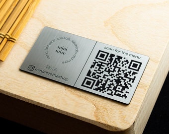 Puede incluir: Una tabla de madera clara con una esterilla de bambú y una placa metálica rectangular. La placa muestra el texto mini mize, un icono de Instagram con el nombre minimizetheshop y un código QR con el texto scan for the menu.
