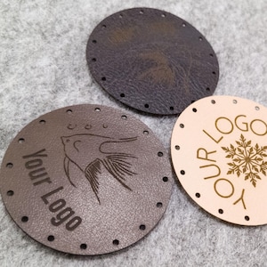 Può includere: Tre etichette rotonde in pelle nelle tonalità del marrone e del beige. Un'etichetta presenta un disegno di pesce e il testo "Your Logo". Un'altra etichetta ha un disegno a fiocco di neve e il testo "Our Logo". Ogni etichetta ha fori distanziati.