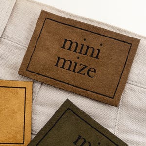 Puede incluir: Primer plano de tres etiquetas rectangulares marrones, beige y verde oliva con la palabra "minimize" impresa en negro. Las etiquetas están sobre una tela beige claro con un patrón de espiga.