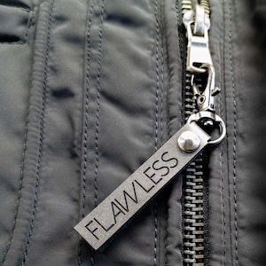 May include: A close-up shot of a dark grey jacket zipper with a silver metal pull. A small, rectangular grey leather tag is attached to the pull, with the word "FLAWLESS" embossed. The jacket is a dark grey colour.