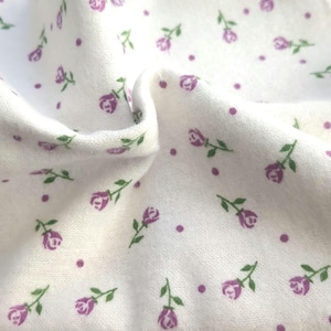 May include: Close-up of white fabric with a repeating pattern of purple roses and dots. The roses have green stems and leaves. The fabric is slightly wrinkled, showing its texture.