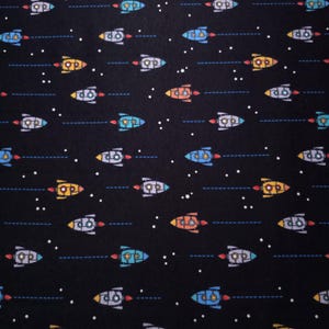 May include: A black fabric featuring a repeating pattern of colorful cartoon rockets in shades of blue, orange, yellow, and grey. Each rocket is depicted with a red exhaust trail and a dashed blue line, interspersed with small white stars.