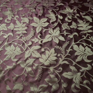 May include: A close-up of a fabric with a repeating pattern of embossed floral designs. The fabric is a deep purple color with a light gold floral pattern.