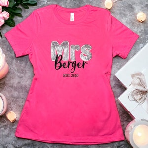 Op de afbeelding: Roze T-shirt met de tekst "Mrs. Berger EST. 2020" in zwarte en zilveren glitterletters.