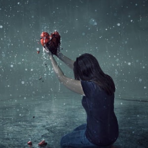 May include: A woman kneels in the rain, holding a broken red heart in her hands. The heart is shattered into pieces, symbolizing heartbreak and sorrow.