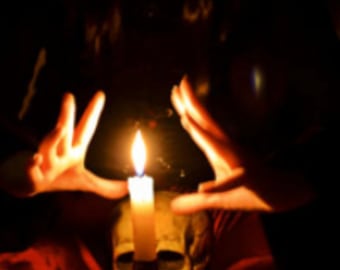 May include: A person's hands are raised in front of a lit candle, which is sitting on top of a skull. The candle is white and the skull is a light brown color. The background is dark.