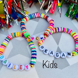 May include: Three colorful beaded bracelets with names spelled out in white letter beads. The bracelets are made of rainbow-colored flat beads and gold accents. The word "Kids" is written in black script below the bracelets.