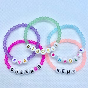 May include: Five pastel-colored beaded bracelets with white letter beads spelling out the names "BIG SIS", "AVA", "QUEEN B", "HADLEY", and "REMY".