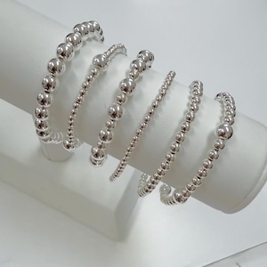 May include: Six silver beaded bracelets of varying sizes are displayed on a white cylindrical stand. The bracelets feature small, round, shiny beads strung together. The beads reflect light, creating a bright and polished appearance. The bracelets are a simple and elegant jewelry item.