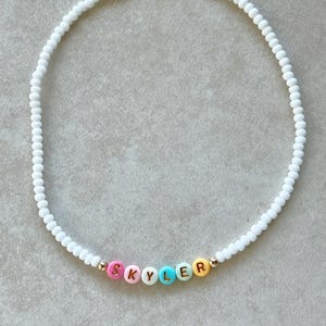 May include: White beaded necklace with colorful letter beads spelling "SKYLER". The letter beads are pink, yellow, blue, and orange. The necklace has a gold-colored clasp.
