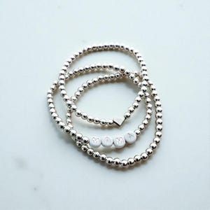 May include: Three silver beaded bracelets. One bracelet features the word "MAMA" spelled out in white letter beads. Another bracelet has a small heart charm. The bracelets are on a white surface.