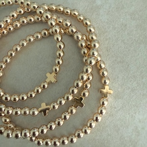 May include: Gold beaded bracelets with small cross charms. The bracelets are made of small, round, gold-colored beads. The crosses are also gold-colored and are evenly spaced along the bracelets. The bracelets are arranged in a layered style.