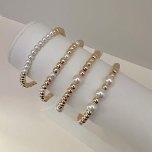 May include: Four bracelets with alternating gold and white beads. The bracelets are stacked on a white display stand.