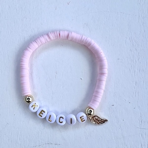 May include: A light pink beaded bracelet with the name "KELCIE" spelled out in white letter beads. Gold-colored beads and a gold wing charm accent the bracelet. The bracelet is made of flat, disc-shaped beads.
