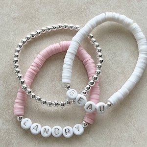 May include: Three beaded bracelets. One bracelet is made of small silver beads. The other two bracelets are made of white and pink disc-shaped beads. The white and pink bracelets have the names "BEAU" and "LANDRY" spelled out in white letter beads.