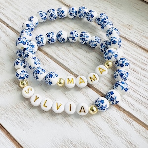 May include: Two stacked bracelets. The top bracelet features white beads with blue floral patterns. The bottom bracelet has white letter beads spelling "Sylvia" and "Mama", interspersed with gold beads. The bracelets are on a light wood surface.