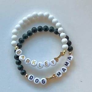 May include: Two beaded bracelets. One bracelet has white beads, the other has black beads. The top bracelet says "BARKLEY" in white letter beads. The bottom bracelet says "DOG MAMA" in white letter beads. Both bracelets have gold bone charms.