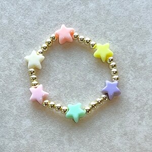 May include: A gold chain bracelet with pastel colored star-shaped beads. The bracelet has a light pink, yellow, white, light green, and purple star bead.