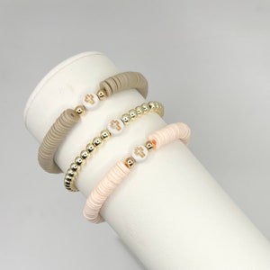 May include: Three bracelets with cross charms. One bracelet is made of beige beads, one is made of gold beads, and one is made of light pink beads.