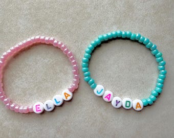 May include: Two beaded bracelets. One is pink with the name "ELLA" spelled out in colorful letter beads. The other is teal with the name "JAYDA" spelled out in colorful letter beads. Both bracelets are on a neutral background.