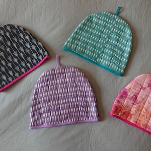 Może przedstawiać: Cztery różne osłonki na czajnik. Jedna jest czarna z białymi wzorami geometrycznymi, druga jest turkusowa z białymi wzorami geometrycznymi, jedna jest fioletowa z białymi wzorami geometrycznymi, a jedna jest różowa z pomarańczowymi i białymi wzorami tie-dye.