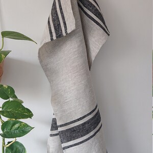 May include: A white linen kitchen towel with black stripes hanging on a wooden hook. The towel is folded and has a natural, rustic look.