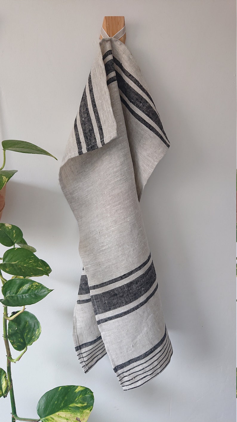 May include: A white linen kitchen towel with black stripes hanging on a wooden hook. The towel is folded and has a natural, rustic look.