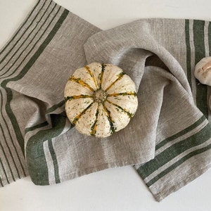 May include: A beige linen kitchen towel with green stripes. The towel is folded and has a small pumpkin and garlic on top.