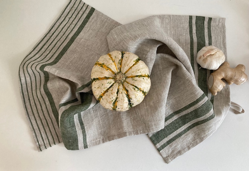 May include: A beige linen kitchen towel with green stripes. The towel is folded and has a small pumpkin and garlic on top.