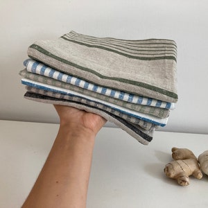May include: A stack of six linen kitchen towels in various patterns. The top towel is a natural linen with green stripes, the second is a blue and white striped towel, the third is a green and white striped towel, the fourth is a blue and white gingham towel, the fifth is a black and white gingham towel, and the bottom towel is a solid black towel.