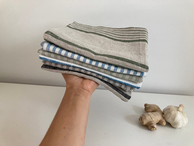 May include: A stack of six linen kitchen towels in various patterns. The top towel is a natural linen with green stripes, the second is a blue and white striped towel, the third is a green and white striped towel, the fourth is a blue and white gingham towel, the fifth is a black and white gingham towel, and the bottom towel is a solid black towel.