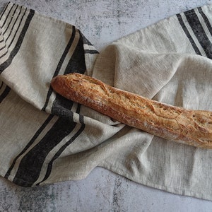 May include: A long, crusty baguette loaf of bread resting on a linen kitchen towel with black stripes.