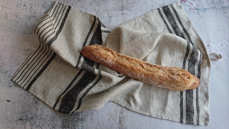 May include: A long, crusty baguette loaf of bread resting on a linen kitchen towel with black stripes.