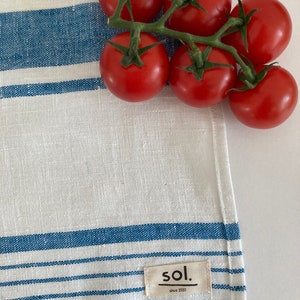 May include: A white linen tea towel with blue stripes and a small tag that says "sol. since 2020" is shown with a cluster of red tomatoes on a vine.