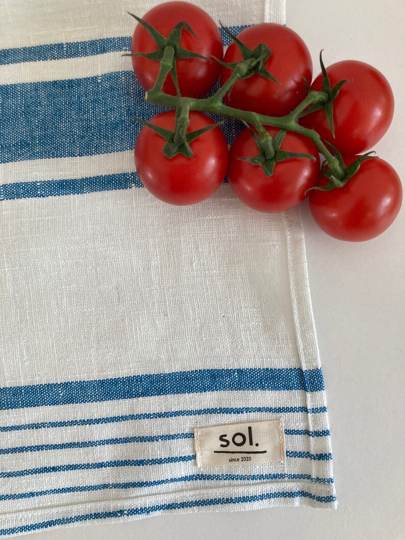 May include: A white linen tea towel with blue stripes and a small tag that says "sol. since 2020" is shown with a cluster of red tomatoes on a vine.