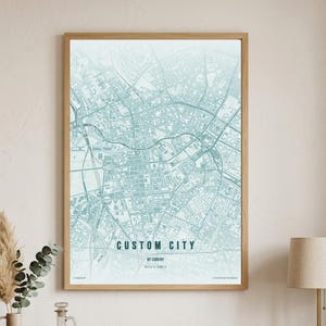 May include: A framed wall art print featuring a light blue and white map of a city grid. The text CUSTOM CITY is printed in black capital letters in the center, with smaller text below. The print is enclosed in a thin, light-colored wooden frame.