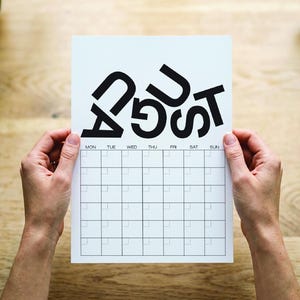 Peut inclure: Une page de calendrier blanche pour août avec une grille de carrés et le nom du mois en grandes lettres noires stylisées. Les jours de la semaine sont imprimés en haut. Le calendrier est tenu sur une surface en bois.