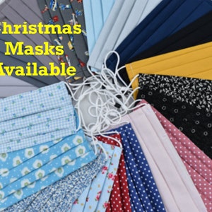 May include: An assortment of fabric face masks in various colours and patterns, including blue floral, polka dots, and solid colours. The text "Christmas Masks Available" is displayed in yellow. Each mask has white elastic ear loops.