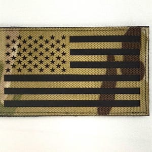 May include: Rectangular patch featuring a stylized American flag design. The flag incorporates a camouflage pattern in shades of tan, brown, and green. The stars and stripes are rendered in solid black, creating a bold contrast. The patch has a sewn border.