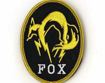 Puede incluir: Parche ovalado negro con un diseño de zorro dorado y la palabra "FOX" en blanco. El zorro está estilizado con rayos. El parche tiene un borde dorado.