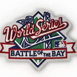 Peut inclure: Écusson brodé avec l'inscription "World Series" en script rouge et blanc. L'écusson comprend le texte "Battle of the Bay" et un logo de baseball. Le design intègre un pont et l'année 1989.