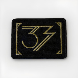 Puede incluir: Un parche rectangular bordado en negro que presenta un diseño cosido en color dorado del número 33. El diseño está encerrado dentro de soportes de esquina dorados, creando una apariencia enmarcada con líneas nítidas y un acabado de tela.