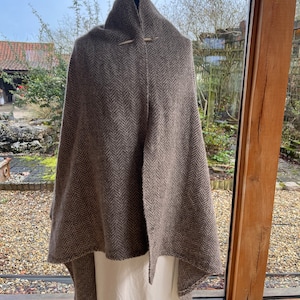 May include: A brown herringbone wool shawl with a wooden pin. The shawl is draped over a white dress form.